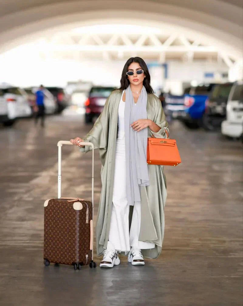 woman wearing flowing bisht-style abaya in pastel green carrying orange handbag and suitcase in parking lot