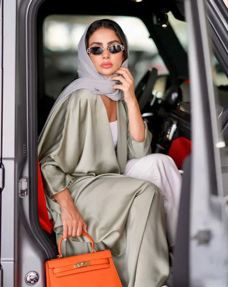 woman wearing flowing bisht-style abaya in pastel green carrying orange handbag and suitcase in parking lot