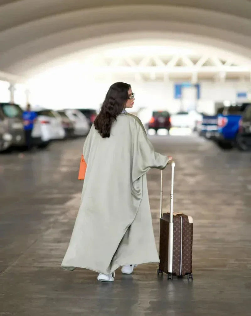 woman wearing flowing bisht-style abaya in pastel green carrying orange handbag and suitcase in parking lot