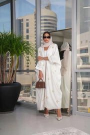 Elegant white linen abaya with gold and white hand-embroidered floral motifs, styled with matching hijab and accessories