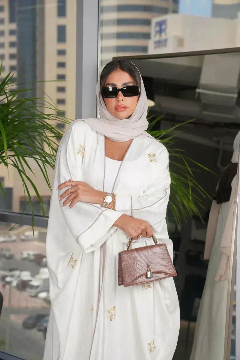Elegant white linen abaya with gold and white hand-embroidered floral motifs, styled with matching hijab and accessories