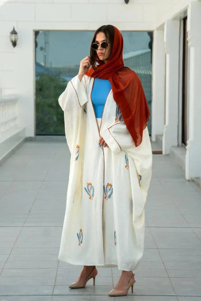 White bisht-style abaya made of natural linen with colorful leaf embroidery, worn with a red hijab and blue top.