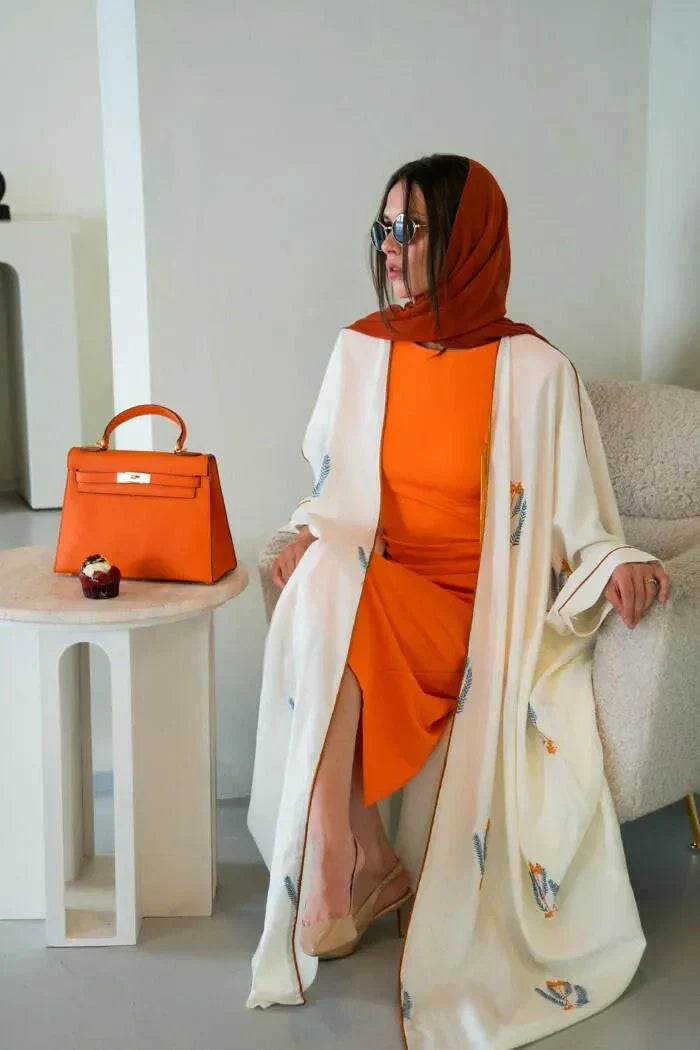 White bisht-style abaya made of natural linen with colorful leaf embroidery, worn with a red hijab and blue top.