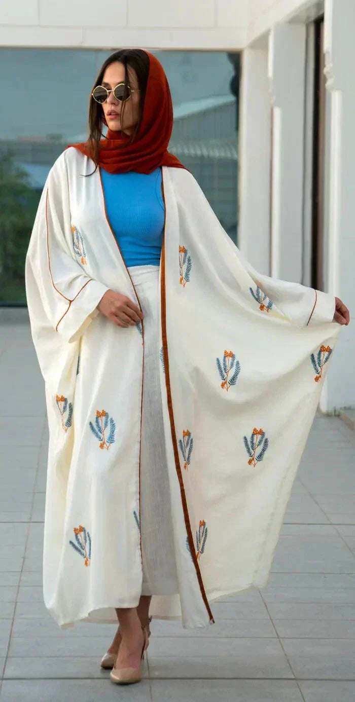 White bisht-style abaya made of natural linen with colorful leaf embroidery, worn with a red hijab and blue top.