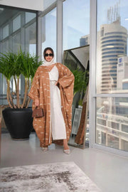 Woman wearing a camel-colored linen bisht with white feather pattern standing indoors by large windows
