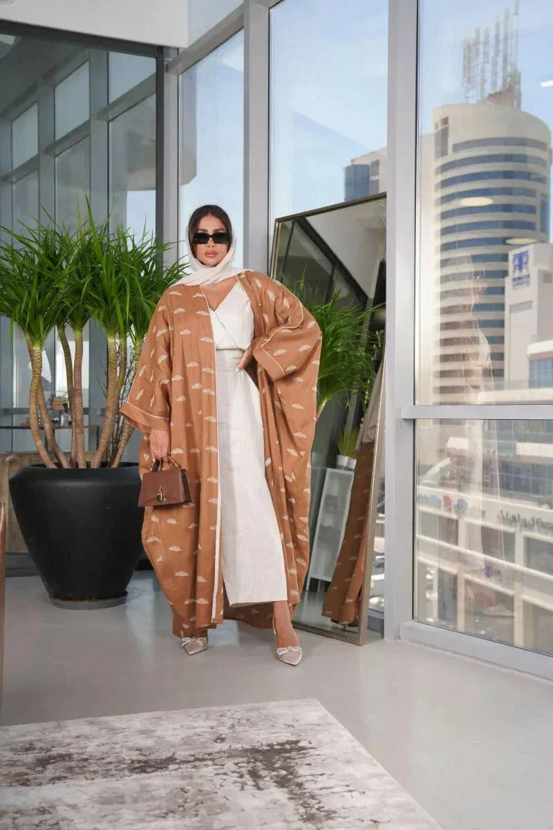 Woman wearing a camel-colored linen bisht with white feather pattern standing indoors by large windows