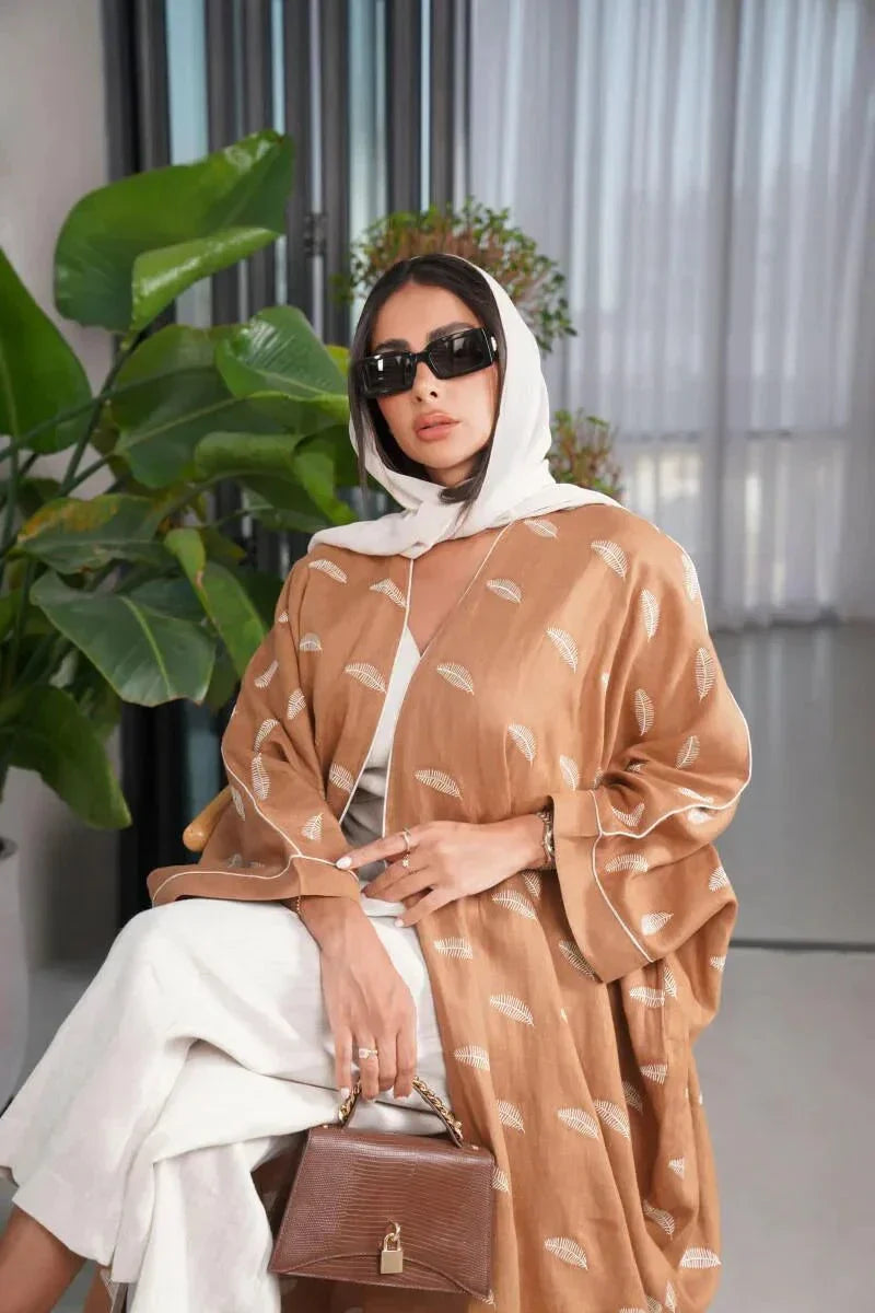 Woman wearing a camel-colored linen bisht with white feather pattern standing indoors by large windows