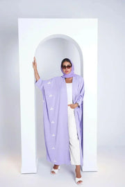 Woman wearing lavender bisht made of soft purple crepe fabric with white clove-inspired embroidery standing in white archway