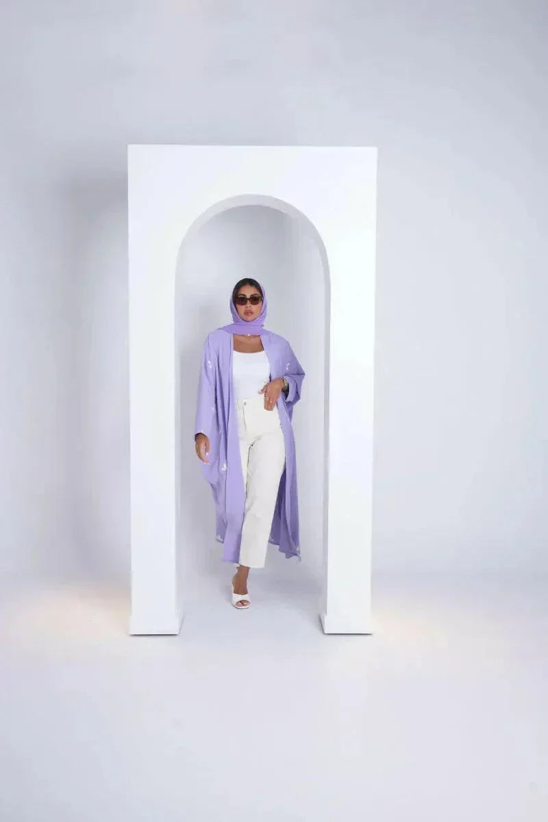 Woman wearing lavender bisht made of soft purple crepe fabric with white clove-inspired embroidery standing in white archway