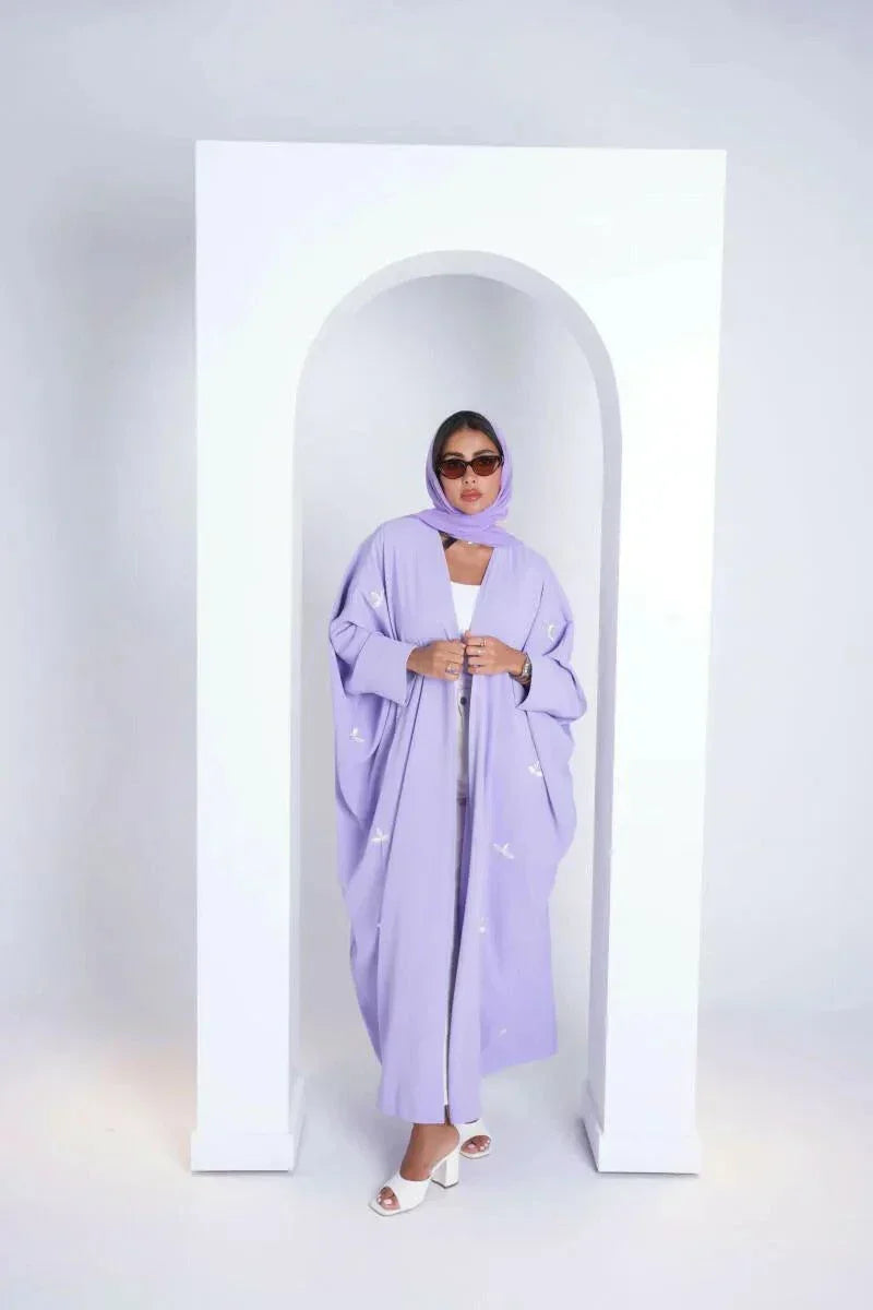 Woman wearing lavender bisht made of soft purple crepe fabric with white clove-inspired embroidery standing in white archway