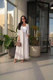 Woman wearing off-white Pearly blazer abaya with side pockets and flowy silhouette, styled with sunglasses and brown handbag indoors.