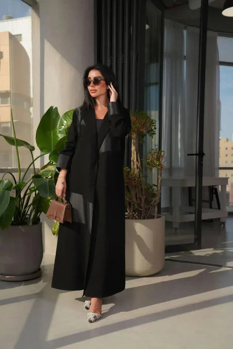 woman wearing modern black abaya with crepe and mokado fabric in a clean blazer silhouette and sharp lapels standing indoors with plants and large windows