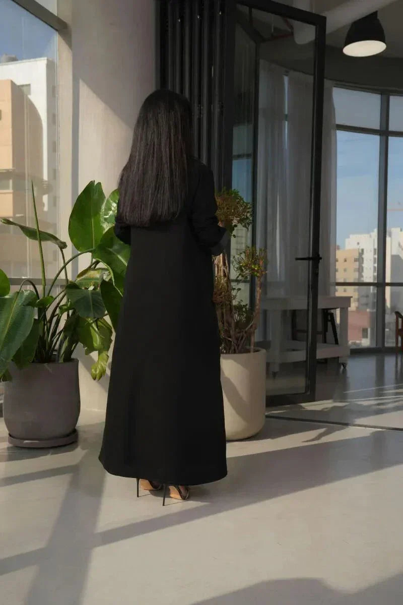 woman wearing modern black abaya with crepe and mokado fabric in a clean blazer silhouette and sharp lapels standing indoors with plants and large windows