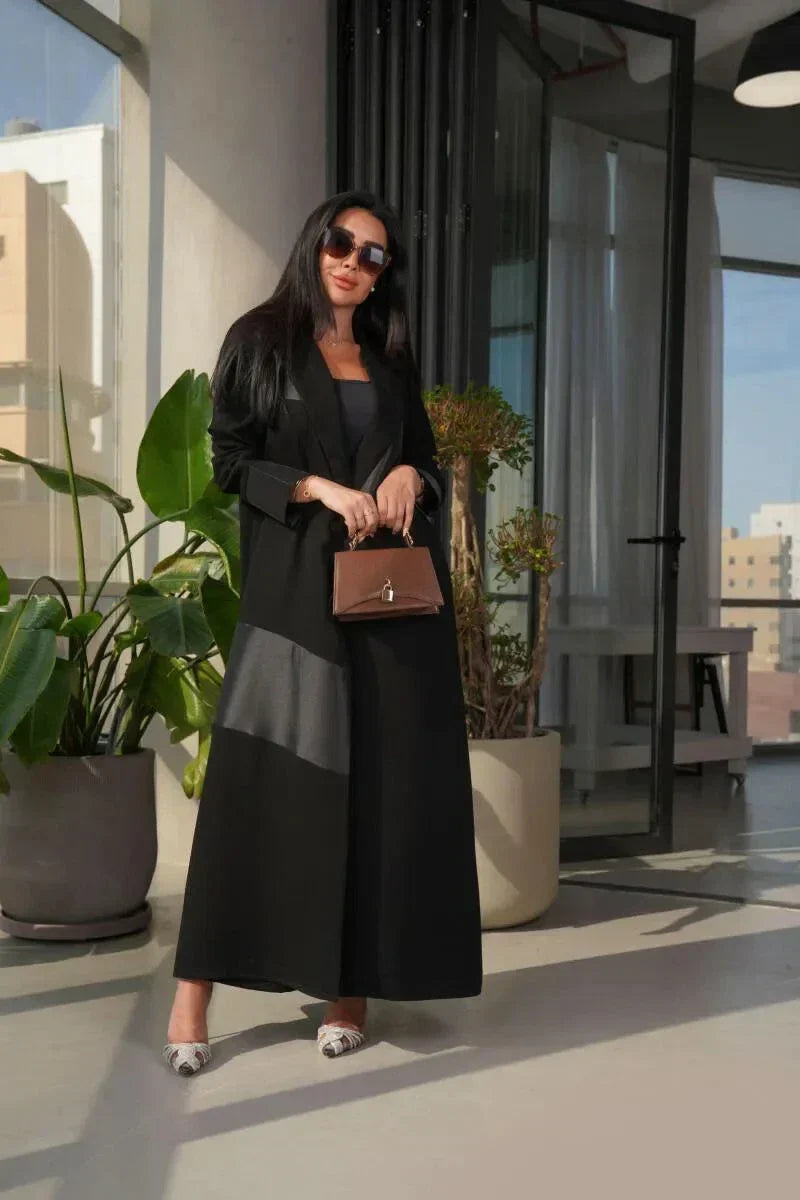 woman wearing modern black abaya with crepe and mokado fabric in a clean blazer silhouette and sharp lapels standing indoors with plants and large windows