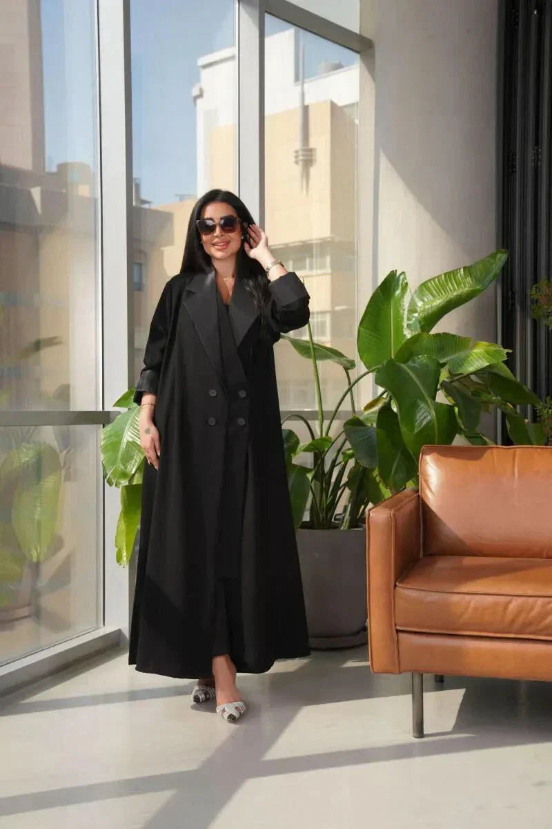 Woman wearing a sleek black blazer-style abaya with double-breasted front and wide lapels standing indoors near plants and a leather chair
