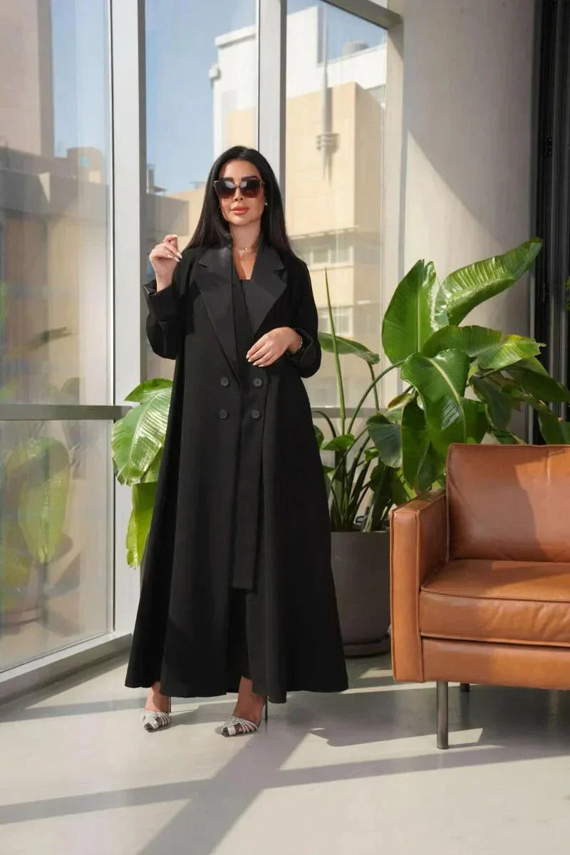 Woman wearing a sleek black blazer-style abaya with double-breasted front and wide lapels standing indoors near plants and a leather chair