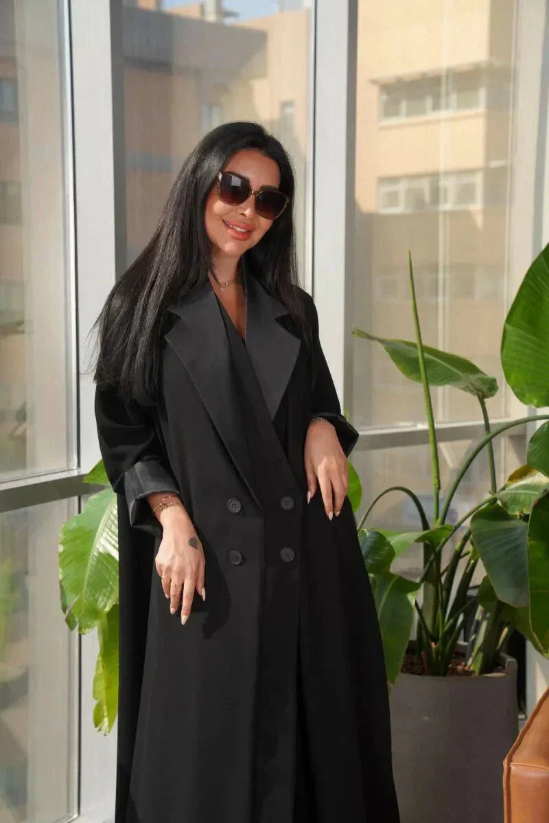 Woman wearing a sleek black blazer-style abaya with double-breasted front and wide lapels standing indoors near plants and a leather chair