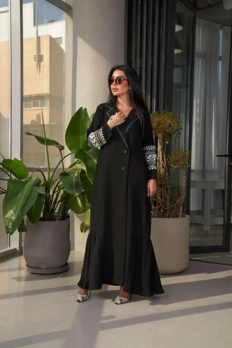 woman wearing a black crepe blazer abaya with white embroidered sleeve cuffs standing indoors near large potted plants