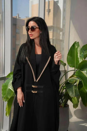 Black crepe abaya with two small leather belts worn by woman in modern indoor setting