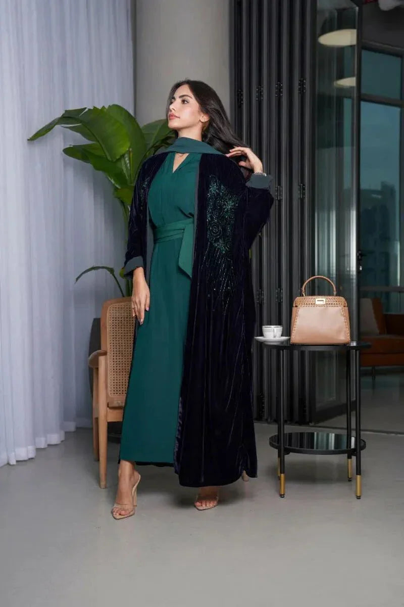 woman wearing luxurious navy blue bisht with green stone embroidery over teal dress standing indoors near chair and table with handbag