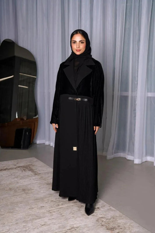 Woman wearing a sleek and modern black abaya with velvet top, pleated crepe skirt, and slim belt in a minimalist indoor setting