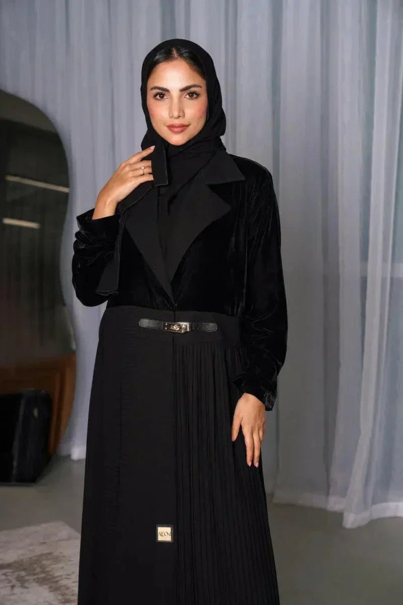 Woman wearing a sleek and modern black abaya with velvet top, pleated crepe skirt, and slim belt in a minimalist indoor setting - sleek and modern abaya
