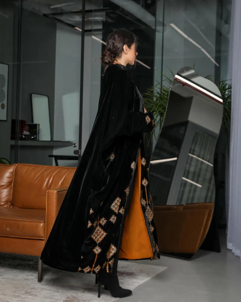 Side view of a woman wearing a luxurious black velvet abaya with intricate gold and beige geometric embroidery and striking orange chiffon lining in a modern indoor setting