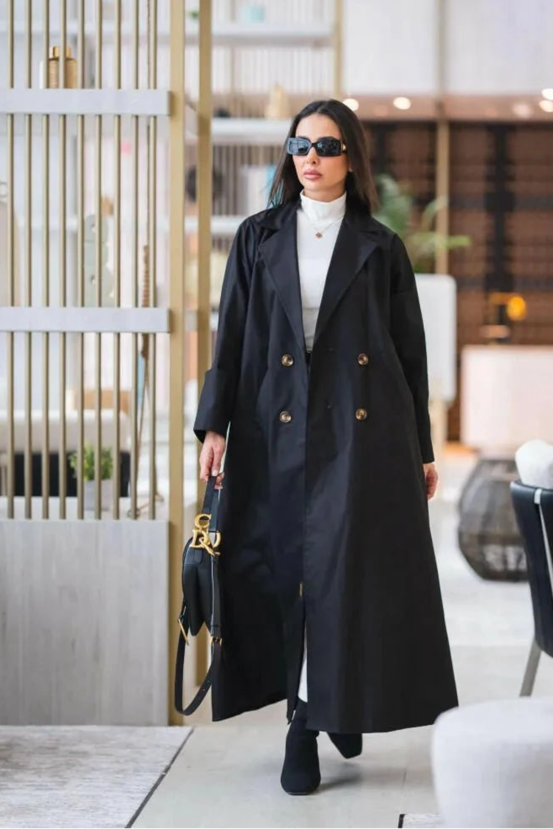 Woman wearing a structured black abaya trench coat with gold buttons and sharp lapels walking indoors