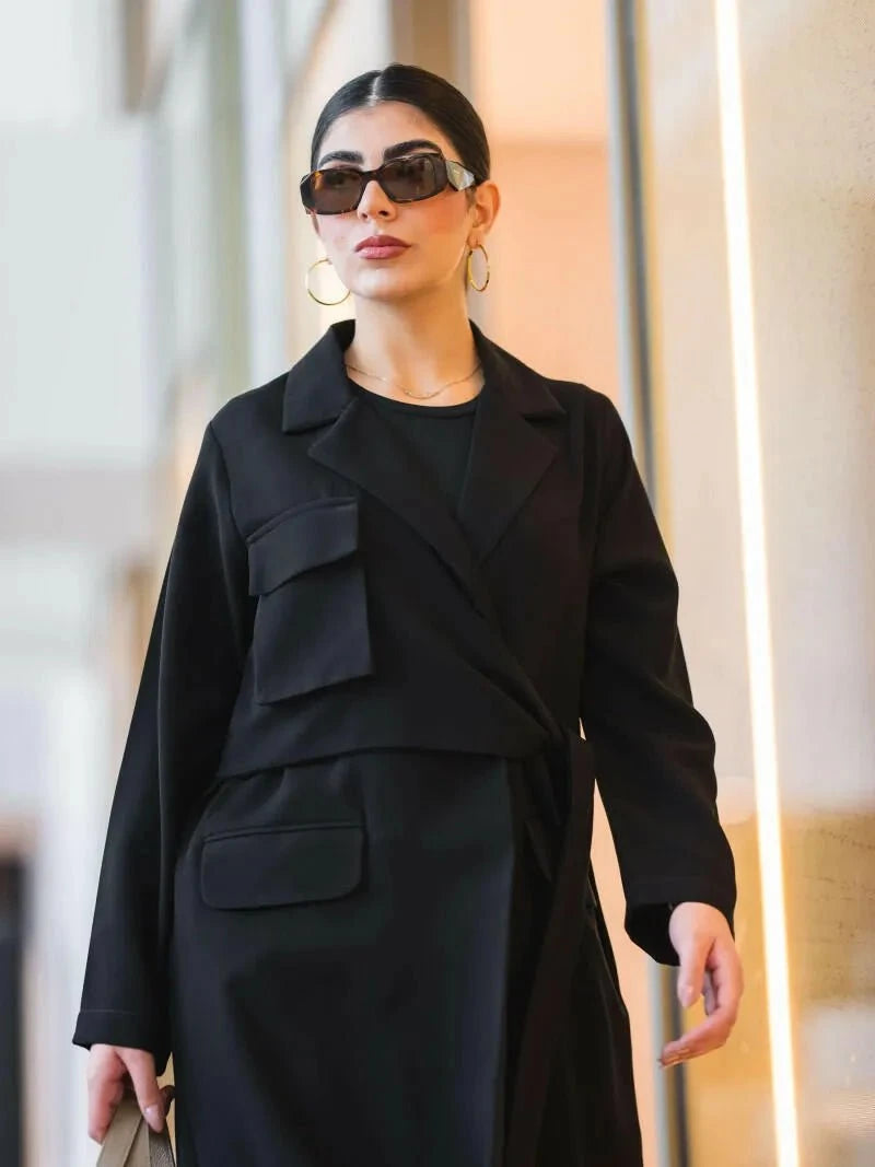 Black blazer-style abaya with structured crepe fabric, sharp lapel collar, oversized flap pockets, and wrap closure.