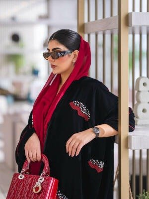 Elegant black velvet abaya with burgundy and white embroidery, paired with a red scarf and handbag.
