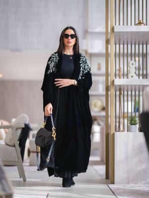 Woman wearing a luxurious black velvet abaya with silver leaf embroidery on shoulders and sleeves, styled with sunglasses and a black handbag.