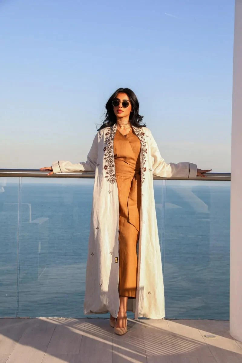 woman wearing white linen abaya with brown geometric embroidery over warm-toned outfit standing by seaside railing