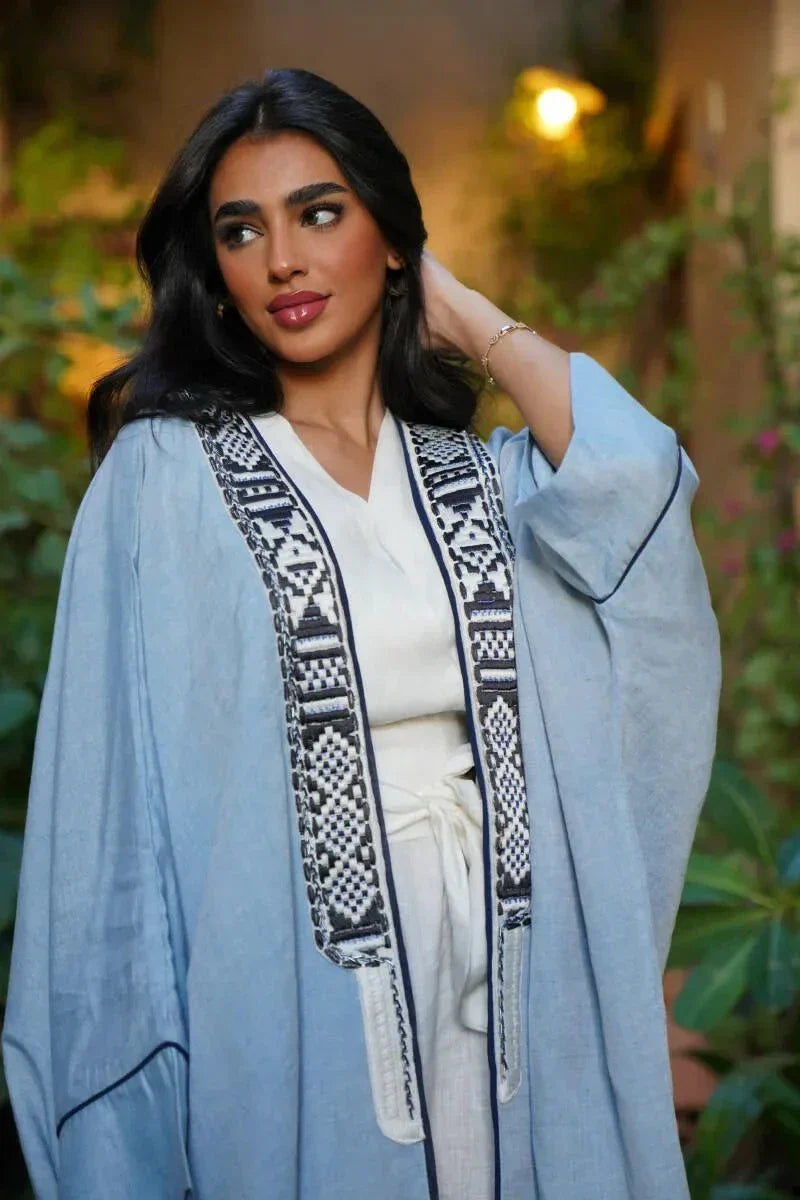 Woman wearing Sahra Blue linen bisht with black-and-white tribal embroidery and wide sleeves over a white dress in a garden setting