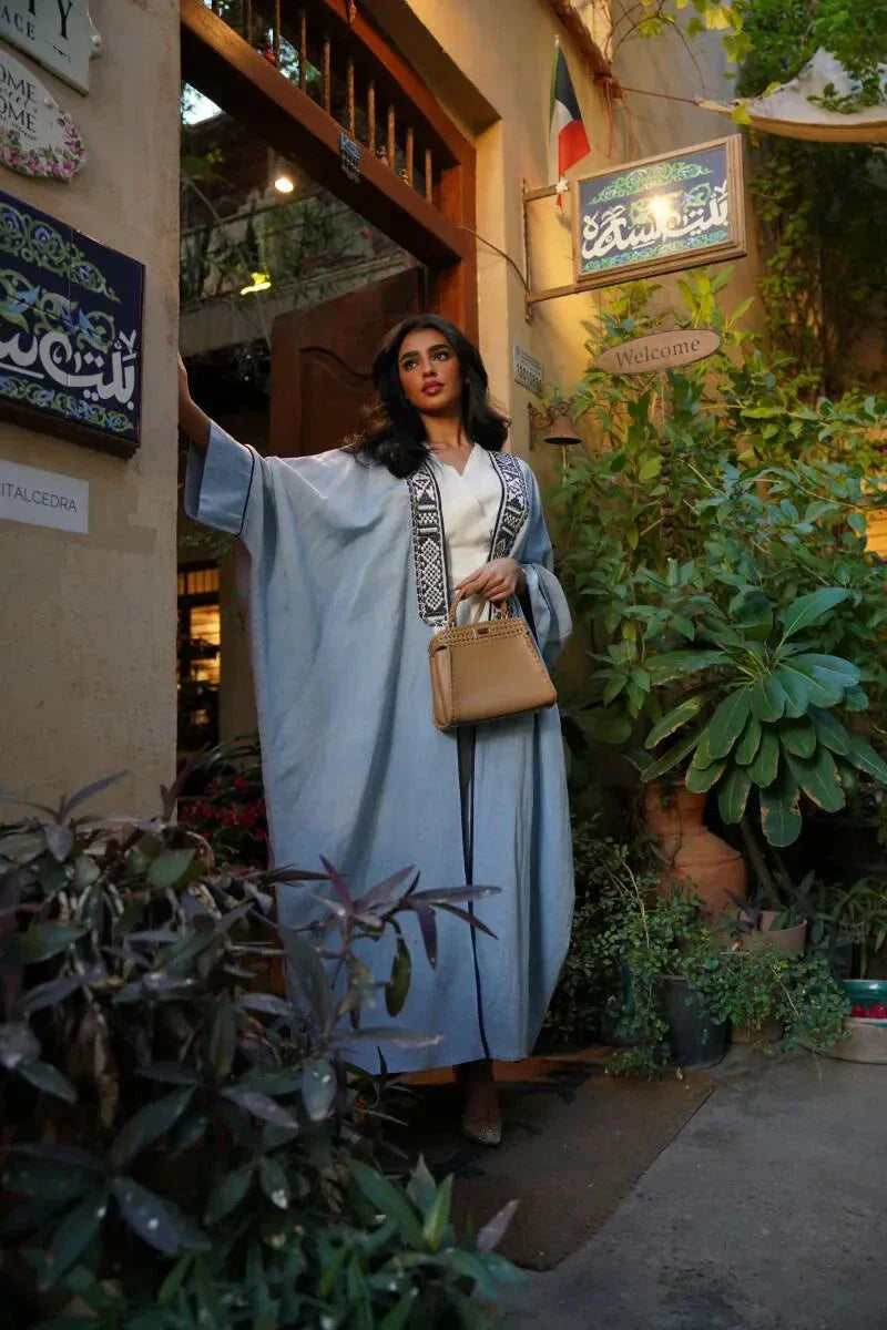 Woman wearing Sahra Blue linen bisht with black-and-white tribal embroidery and wide sleeves over a white dress in a garden setting