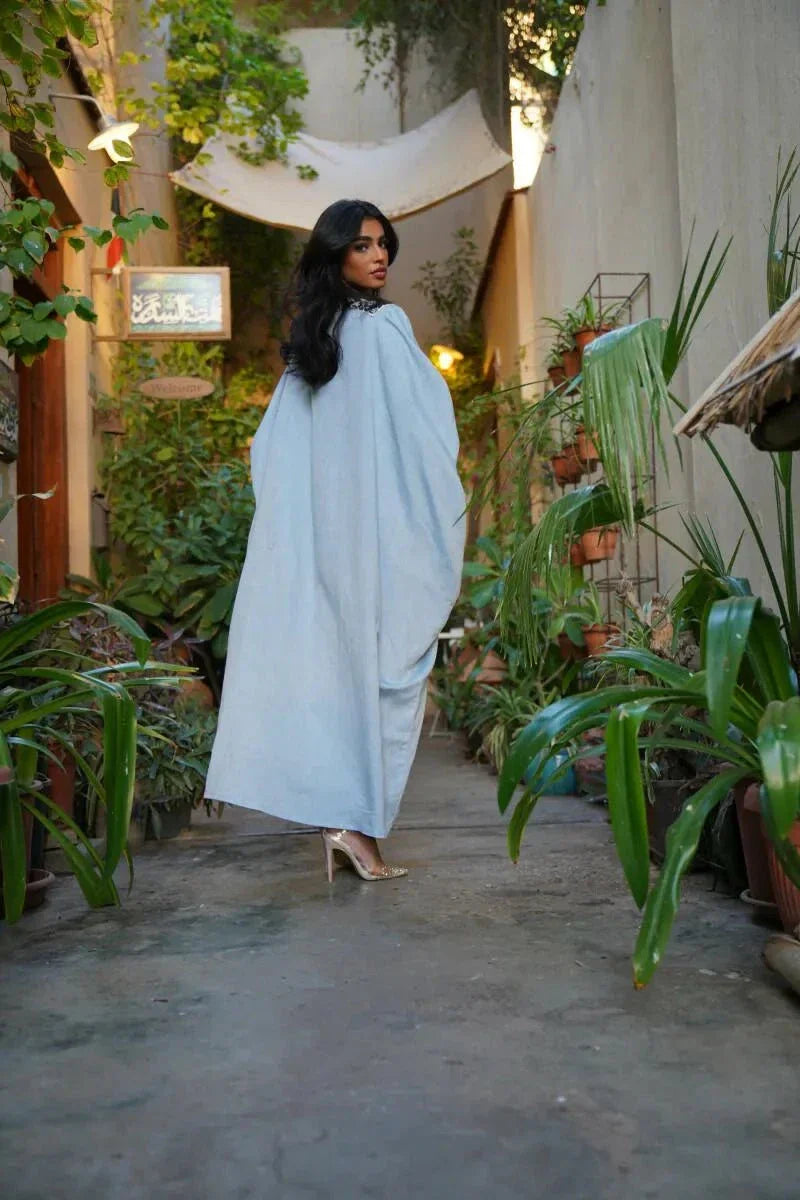 Woman wearing Sahra Blue linen bisht with black-and-white tribal embroidery and wide sleeves over a white dress in a garden setting