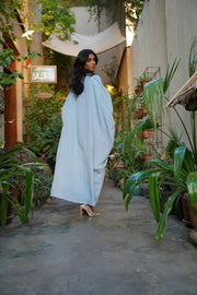 Woman wearing Sahra Blue linen bisht with black-and-white tribal embroidery and wide sleeves over a white dress in a garden setting