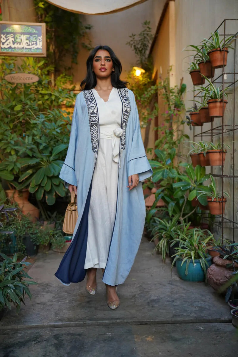 Woman wearing Sahra Blue breathable linen bisht with black-and-white tribal embroidery and wide sleeves walking outdoors surrounded by plants