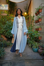 Woman wearing Sahra Blue breathable linen bisht with black-and-white tribal embroidery and wide sleeves walking outdoors surrounded by plants