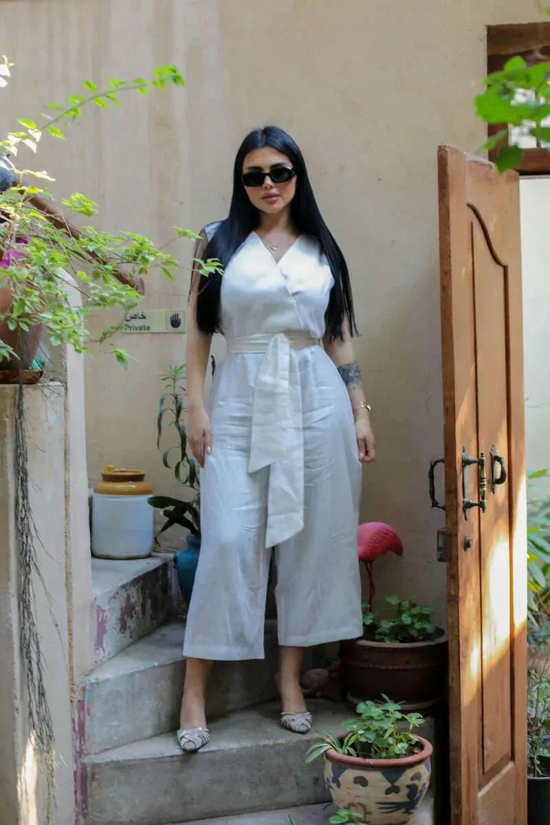 Woman wearing a sleeveless white linen jumpsuit with wrap V-neck and wide belt, standing outdoors with plants - White Jumpsuit Linen