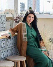 Woman wearing camel-brown linen abaya with green and gold embroidery and piping, styled over a green outfit, sitting outdoors - Honeywood
