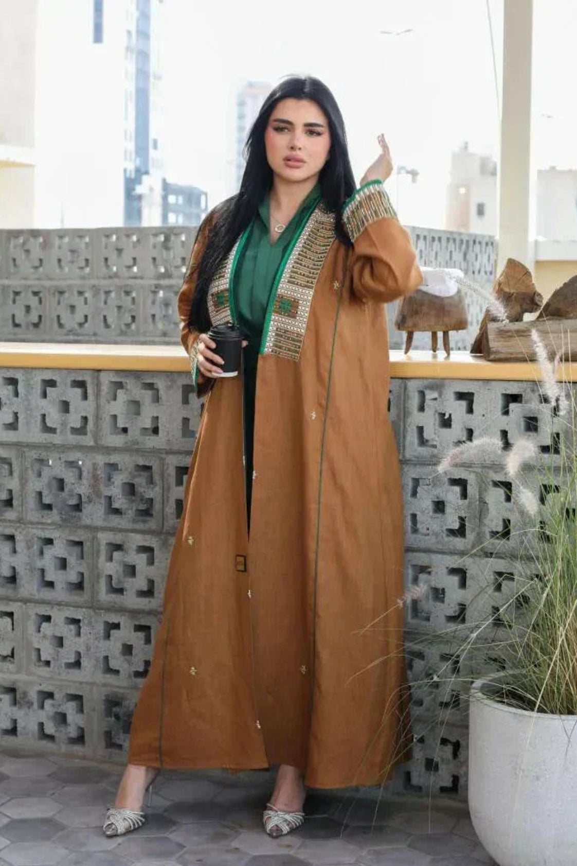 Woman wearing camel-brown linen abaya with green and gold embroidery and piping, styled over a green outfit, sitting outdoors - Honeywood
