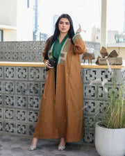 Woman wearing camel-brown linen abaya with green and gold embroidery and piping, styled over a green outfit, sitting outdoors - Honeywood