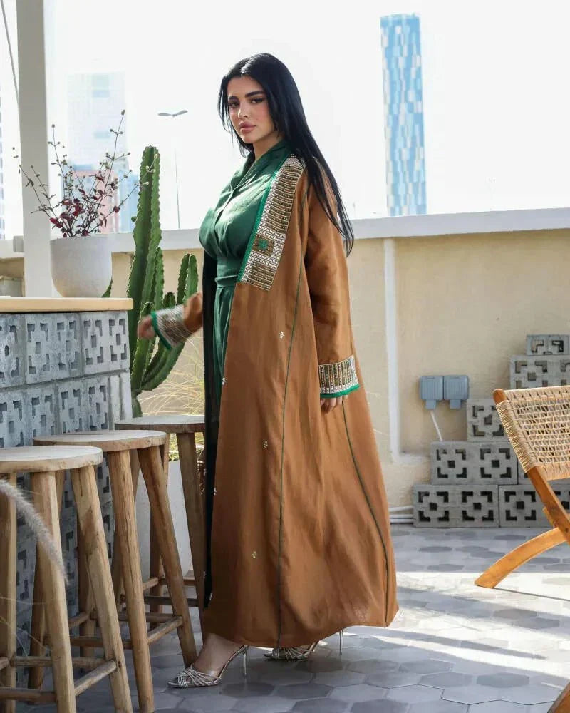 Woman wearing camel-brown linen abaya with green and gold embroidery and piping, styled over a green outfit, sitting outdoors - Honeywood