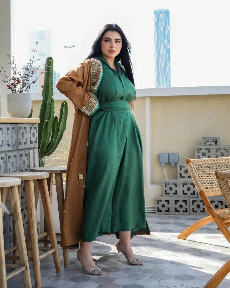 Woman wearing a sleek green linen jumpsuit with a relaxed straight-cut silhouette and V-neckline, styled with a long brown coat on a rooftop terrace.