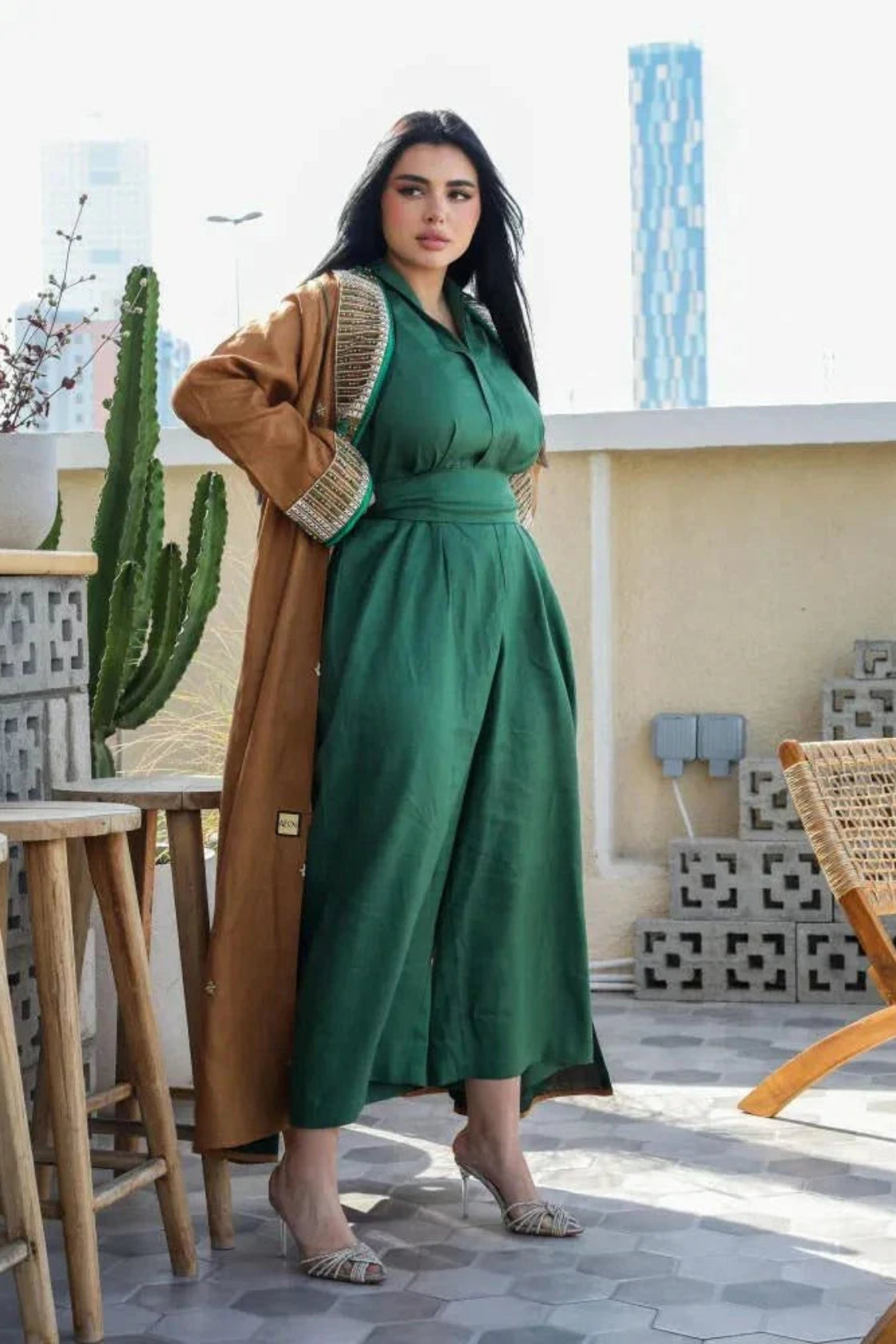 Woman wearing a sleek green linen jumpsuit with a relaxed straight-cut silhouette and V-neckline, styled with a long brown coat on a rooftop terrace.