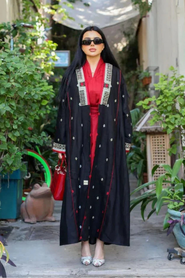 Woman wearing a black linen abaya with pearl embroidery and red piping over a crimson inner dress, accessorized with sunglasses and a red handbag, standing outdoors.