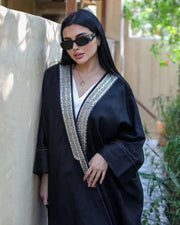 Woman wearing black linen bisht with gold embroidery over white outfit standing outdoors