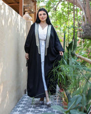Woman wearing black linen bisht with gold embroidery over white outfit standing outdoors