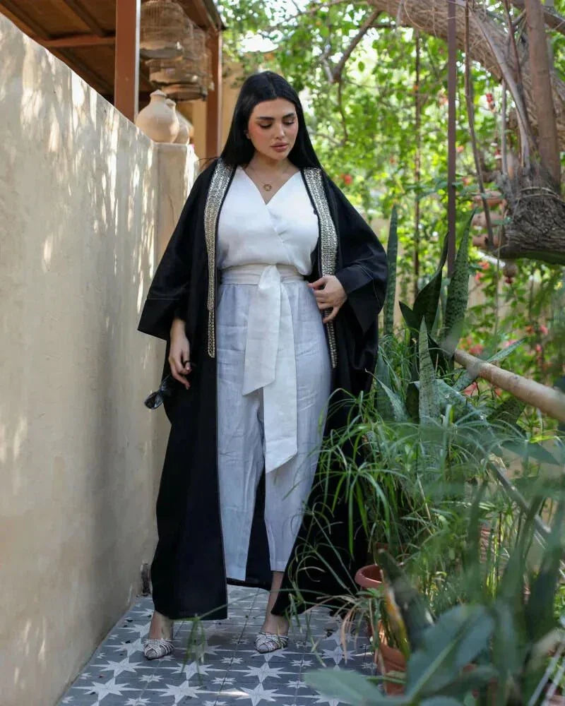 Woman wearing black linen bisht with gold embroidery over white outfit standing outdoors surrounded by plants