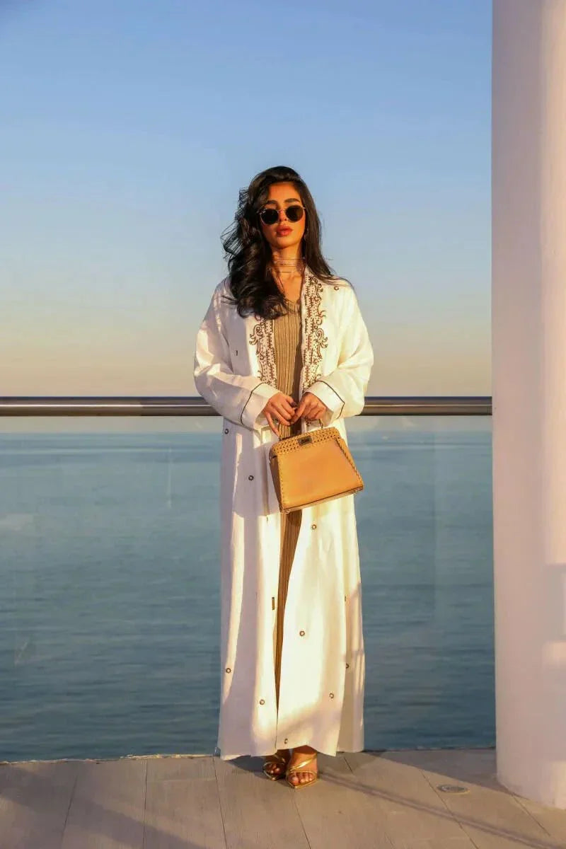 Woman wearing elegant white linen abaya with beige leaf embroidery standing by the sea holding a tan handbag.